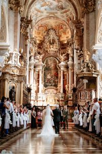 Bride walking down the aisle with her father, photographed from behind, with the impressive altar of Stiftskirche Klosterneuburg in Vienna in view
