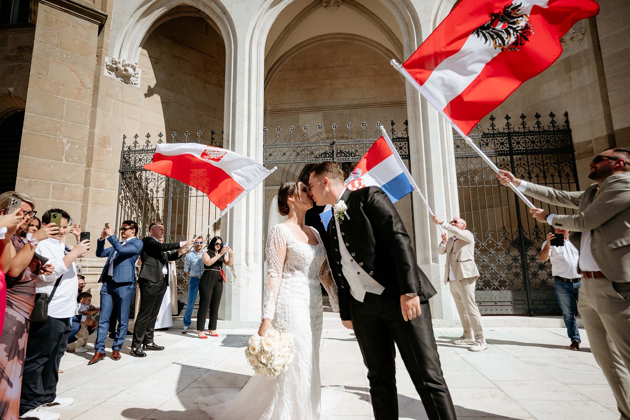 Bride and groom kissing in front of Stiftskirche Klosterneuburg while guests wave Polish and Austrian flags.