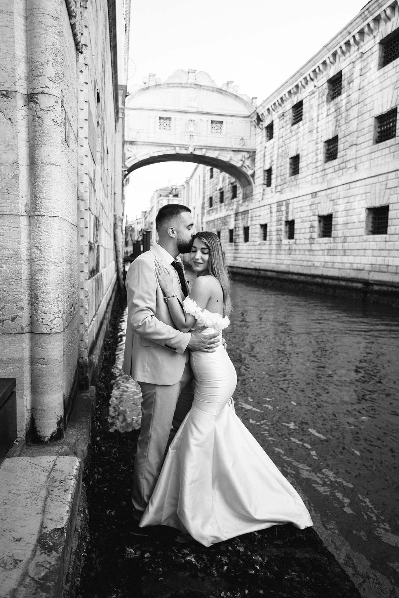 Bride and groom embracing by a Venetian canal, with the groom kissing the bride’s forehead and the Bridge of Sighs in the background.